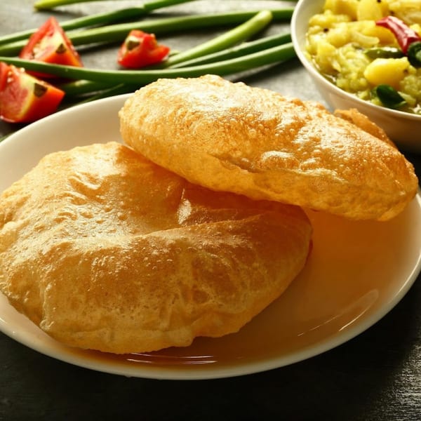 Two golden-brown Indian puri breads on a white plate, with a bowl of potato curry and vegetables in the background.
